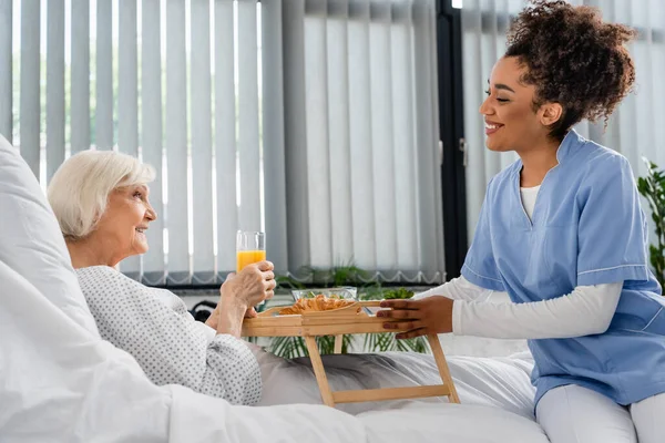 Vista lateral da bandeja de seguradora afro-americana sorridente com alimentos perto do paciente com suco de laranja na enfermaria do hospital — Fotografia de Stock