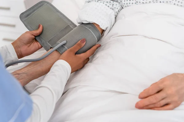 Vista recortada de la enfermera afroamericana usando tonómetro en la mano del paciente anciano en la cama de hospital - foto de stock