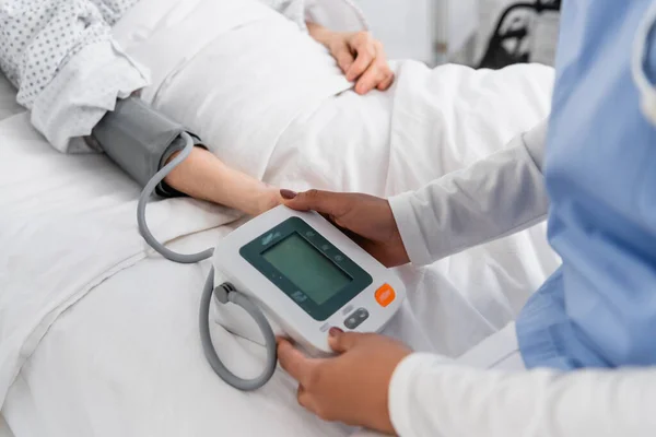 Vista recortada de enfermera afroamericana usando tonómetro digital cerca de paciente en cama de hospital - foto de stock