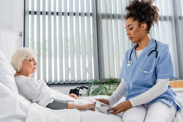 Бічний вид на американську медсестру з використанням тонометра біля літньої пацієнтки в клініці. — стокове фото