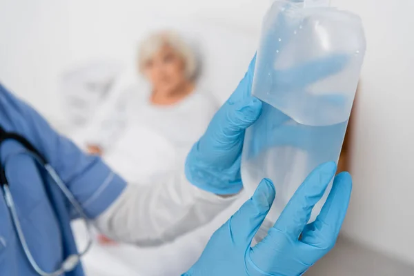 Krankenschwester in Latexhandschuhen hält Flasche mit intravenöser Therapie in der Nähe verschwommener Patientin — Stockfoto