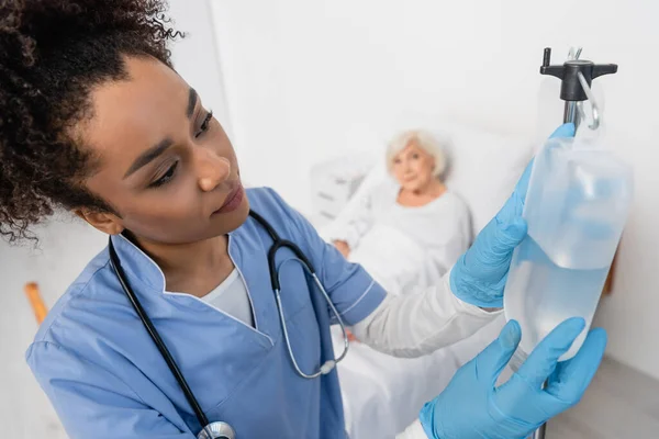 Enfermera afroamericana en guantes de látex sosteniendo botella de terapia intravenosa cerca de paciente borroso en la sala de hospital - foto de stock