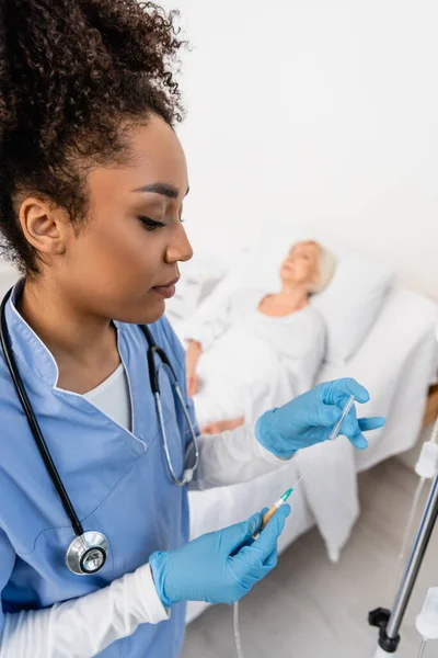 Enfermera afroamericana en guantes de látex sosteniendo catéter cerca de estación de terapia intravenosa en hospital - foto de stock