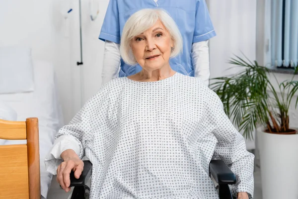 Femme âgée en fauteuil roulant regardant la caméra près de l'infirmière à la clinique — Photo de stock