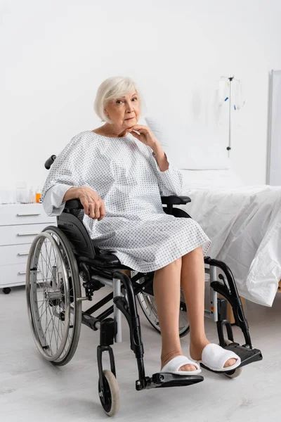 Paciente anciano en bata mirando la cámara en silla de ruedas en la clínica - foto de stock