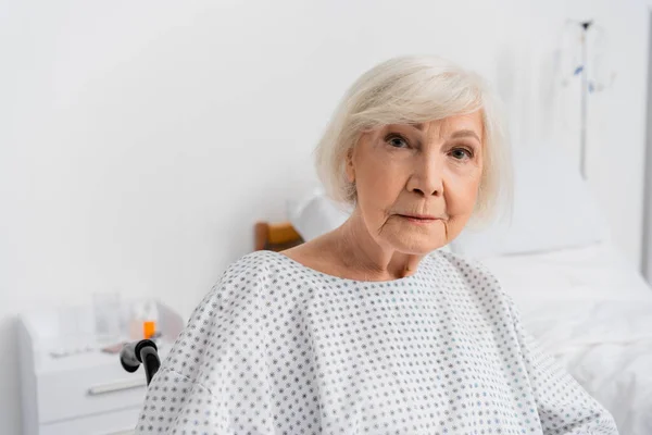 Mujer mayor en bata de paciente mirando a la cámara en silla de ruedas - foto de stock