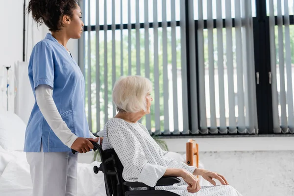 Seitenansicht einer afrikanisch-amerikanischen Krankenschwester in Uniform, die neben einem Patienten im Rollstuhl im Krankenhaus steht — Stockfoto