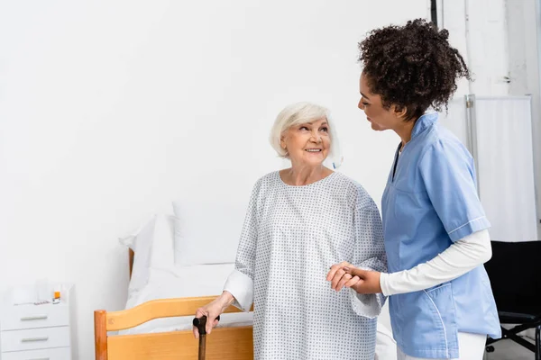 Cheerful elderly woman with walking cane holding hand of african american nurse in hospital ward — Stock Photo