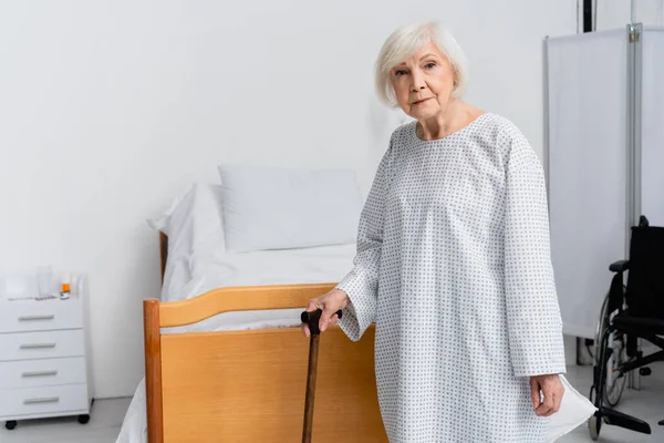 Paciente idoso com bengala andando olhando para a câmera perto da cama e cadeira de rodas — Fotografia de Stock