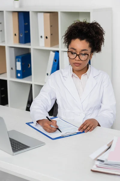 Médecin afro-américain dans les lunettes d'écriture sur presse-papiers près d'un ordinateur portable flou — Photo de stock
