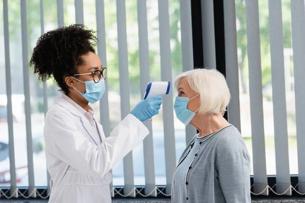 Médico afroamericano en máscara médica comprobando la temperatura del paciente con pirómetro - foto de stock