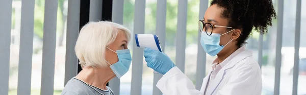 African american doctor in medical mask holding pyrometer near senior patient, banner — Stock Photo