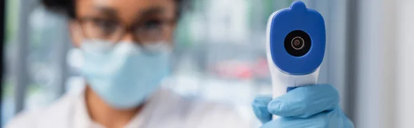 Pyrometer in hand of african american doctor on blurred background, banner — Stock Photo