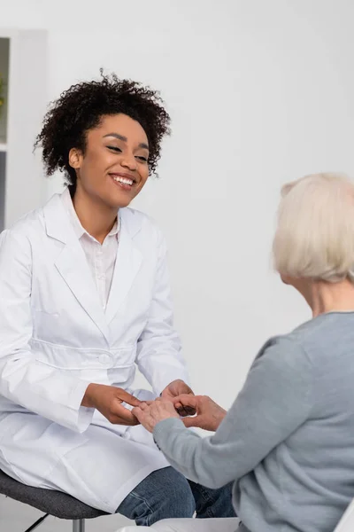 Smiling african american doctor holding hands of senior patient on blurred foreground — Stock Photo