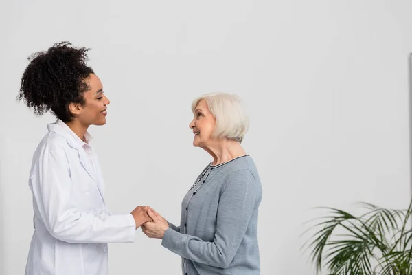 Seitenansicht einer fröhlichen Seniorin, die einem afrikanisch-amerikanischen Arzt in der Klinik die Hand hält — Stockfoto