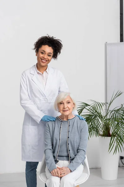 Sorridente medico africano americano in guanti di lattice che abbraccia il paziente anziano — Foto stock