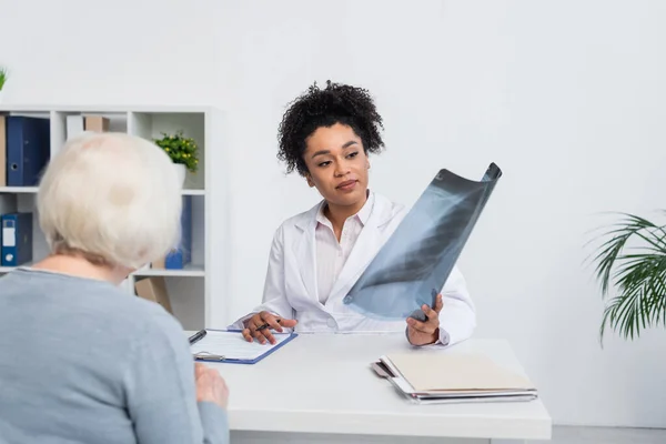 Afrikanisch-amerikanischer Arzt hält Fluorographie in der Nähe von Seniorin und Klemmbrett in Klinik — Stockfoto