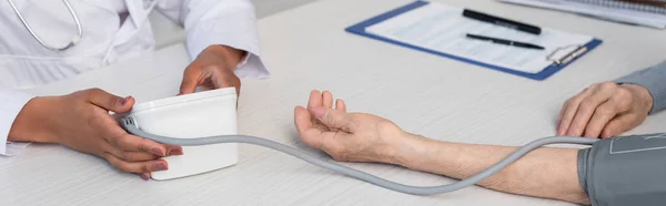 Ausgeschnittene Ansicht eines afrikanisch-amerikanischen Arztes mit Tonometer in der Nähe eines Patienten in der Klinik, Banner — Stockfoto