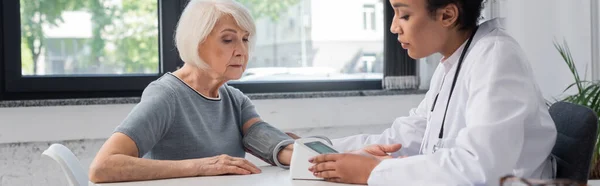 Médecin afro-américain en manteau blanc regardant un tonomètre près d'un patient âgé à l'hôpital, bannière — Photo de stock