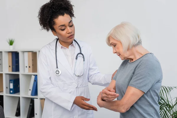 Médico afroamericano mirando el brazo de una mujer mayor en la clínica - foto de stock