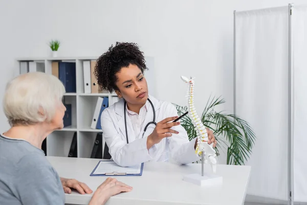 Junger afrikanisch-amerikanischer Arzt zeigt auf verschwommenem Vordergrund auf Wirbelsäulenmodell in der Nähe eines älteren Patienten — Stockfoto