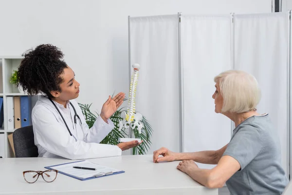 Fisioterapeuta afro-americano apontando para o modelo da coluna vertebral enquanto conversa com paciente sênior — Fotografia de Stock