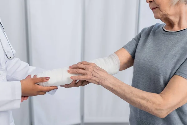 Vista recortada de la mujer mayor con vendaje de yeso en el brazo de pie cerca del médico afroamericano - foto de stock