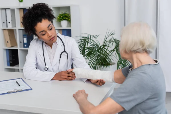 Afro-américain traumatologue vérifier bras du patient âgé en bandage de plâtre — Photo de stock