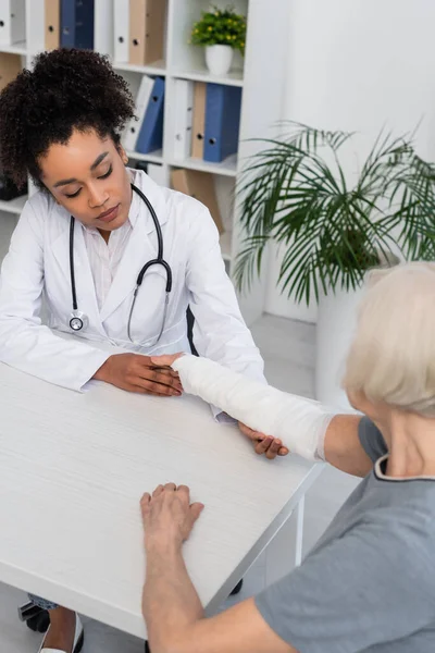 African american doctor looking at arm of patient in plaster bandage — Stock Photo