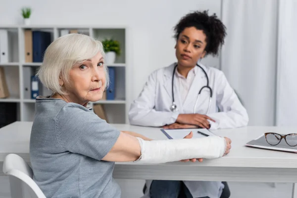 Donna anziana con bendaggio in gesso sul braccio guardando la fotocamera vicino offuscata medico afroamericano — Foto stock