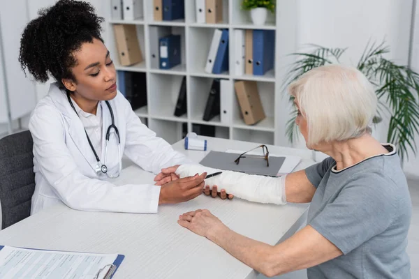 Médico afroamericano señalando vendaje de yeso en el brazo del paciente - foto de stock