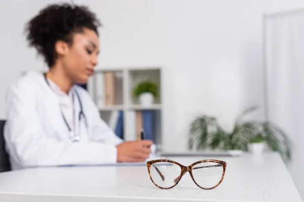 Eyeglasses on table near blurred african american doctor — Stock Photo