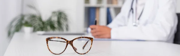 Cropped view of eyeglasses on table near blurred doctor in hospital, banner — Stock Photo