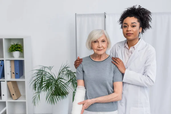 Medico afroamericano che abbraccia paziente con bendaggio in gesso sul braccio — Foto stock