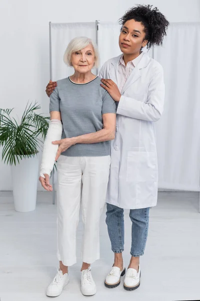 African american doctor hugging patient with plaster bandage on broken arm in hospital — Stock Photo