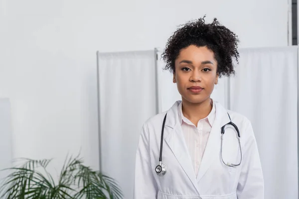 Joven médico afroamericano en bata blanca y estetoscopio mirando la cámara en la clínica - foto de stock