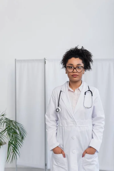 Médico afroamericano con anteojos tomados de la mano en bolsillos de abrigo blanco - foto de stock