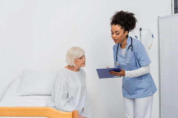 Infirmière afro-américaine avec presse-papiers debout près du patient en robe — Photo de stock