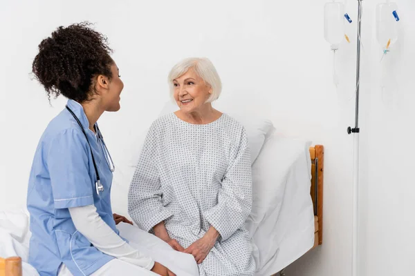 Senior Patient blickt auf afrikanisch-amerikanische Krankenschwester auf Krankenhausbett — Stockfoto