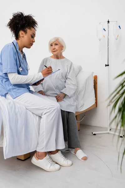 Afrikanisch-amerikanische Krankenschwester schreibt auf Klemmbrett in der Nähe von älteren Patienten auf Krankenhausstation — Stockfoto