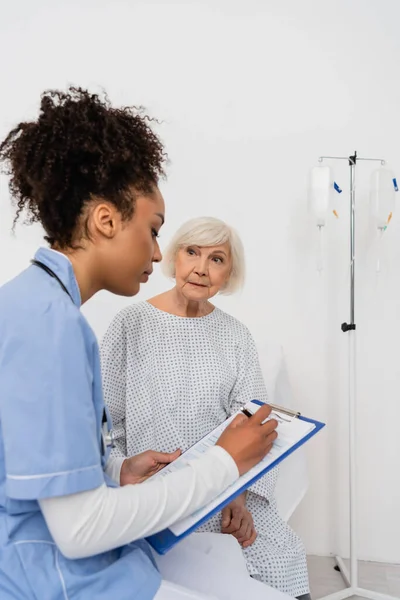 Senior patient looking at african american nurse writing on clipboard — Stock Photo