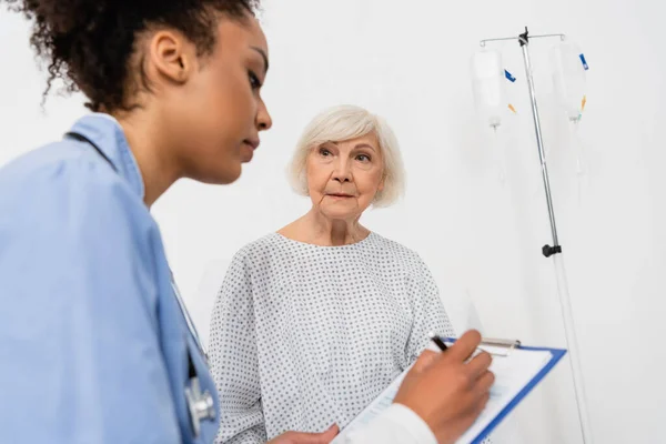 Ältere Patientin im Kleid betrachtet verschwommene afrikanische Krankenschwester, die auf Klemmbrett schreibt — Stockfoto