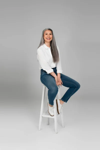 Alegre asiático mulher no azul jeans e branco camisa sentado no alto banquinho no cinza fundo — Fotografia de Stock