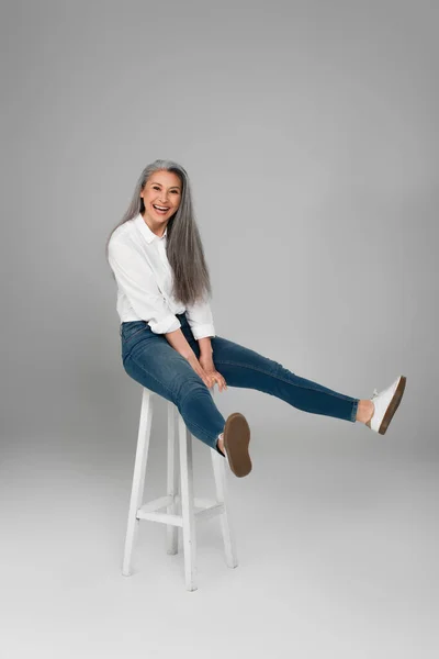 Mature asian woman in blue jeans and white shirt laughing on high stool on grey background — Stock Photo
