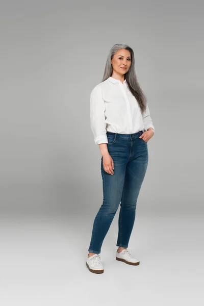 Piena vista lunghezza di donna asiatica sorridente in jeans blu e camicia bianca su grigio — Foto stock