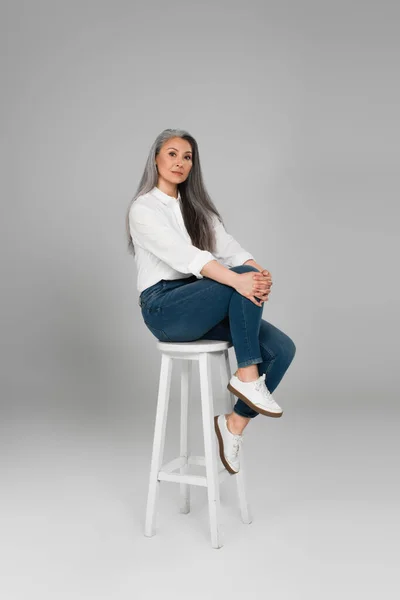 Grey haired asian woman in white shirt and blue jeans looking at camera on high stool on grey background — Stock Photo