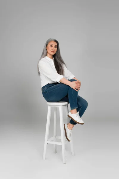 Gris peludo asiático mujer en blanco camisa y azul jeans mirando cámara en alto taburete en gris fondo - foto de stock