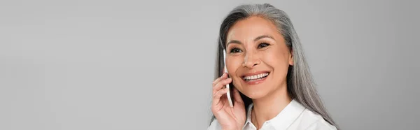 Felice donna asiatica con i capelli grigi che chiama sul cellulare isolato su grigio, banner — Foto stock