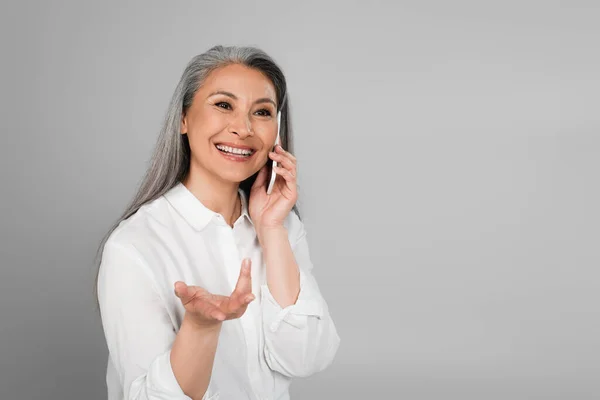 Mature asian woman in white shirt smiling and gesturing while talking on mobile phone isolated on grey — Stock Photo