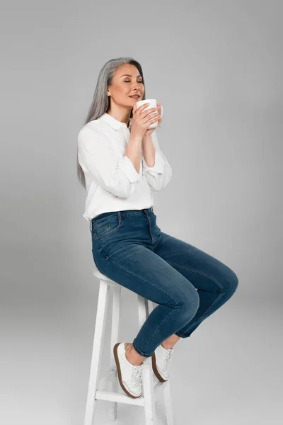 Middle aged asian woman in white shirt and jeans sitting on stool with cup of aromatic tea on grey — Stock Photo